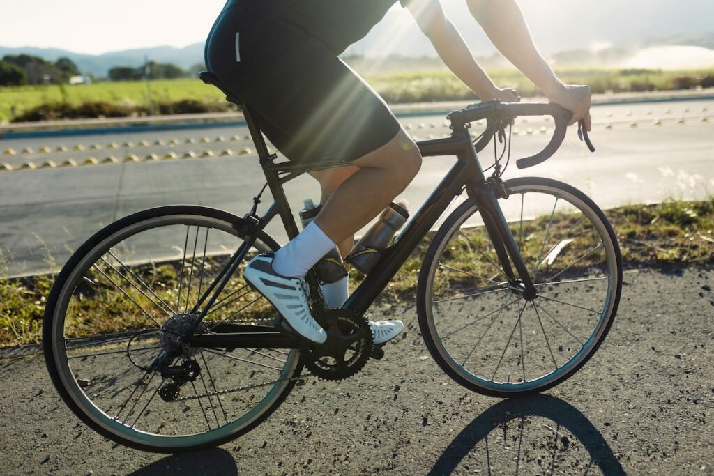 Cyclist in dark shorts pedaling a road bike on a sunlit road, close-up on legs and pedal stroke, demonstrating glute and leg engagement while cycling
