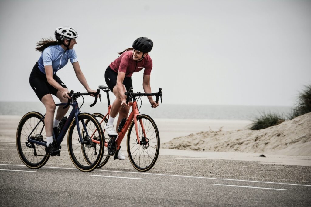 Two women road cycling side by side along a coastal path, standing out of the saddle while pedaling to build glutes and leg strength