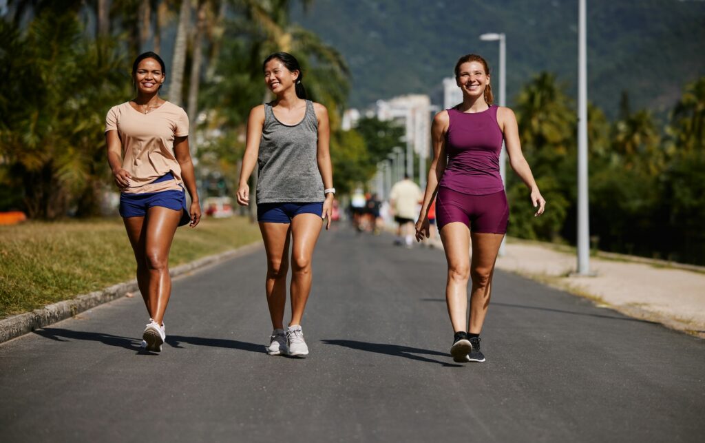 Three women walking briskly outdoors on a tree-lined path, exploring whether does walking build muscle when paired with pace and terrain changes