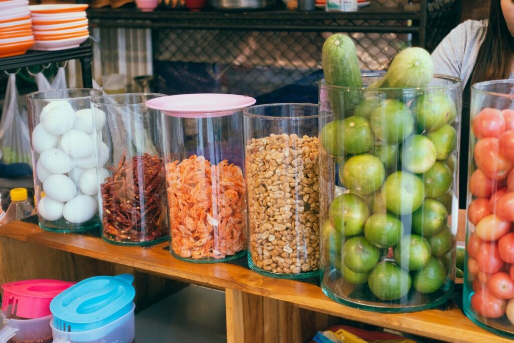 Glass storage jars neatly organized on a wooden shelf, each filled with a single ingredient like eggs, dried chilies, peanuts, limes, and tomatoes, showing an example of clutter free kitchen organization.