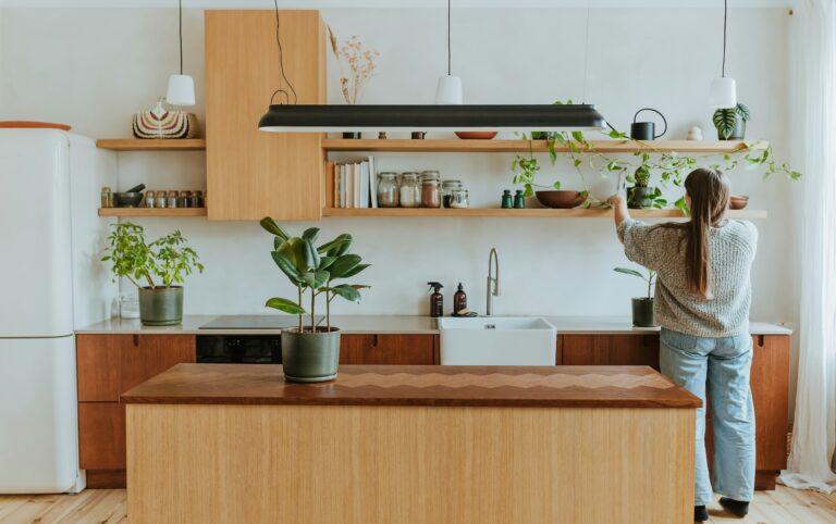 Woman organizing open wooden shelves in a bright kitchen with jars, plants, and books, decluttering and tidying up her kitchen space.