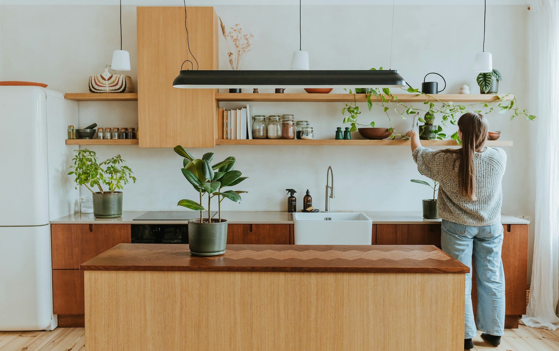 Woman organizing open wooden shelves in a bright kitchen with jars, plants, and books, decluttering and tidying up her kitchen space.