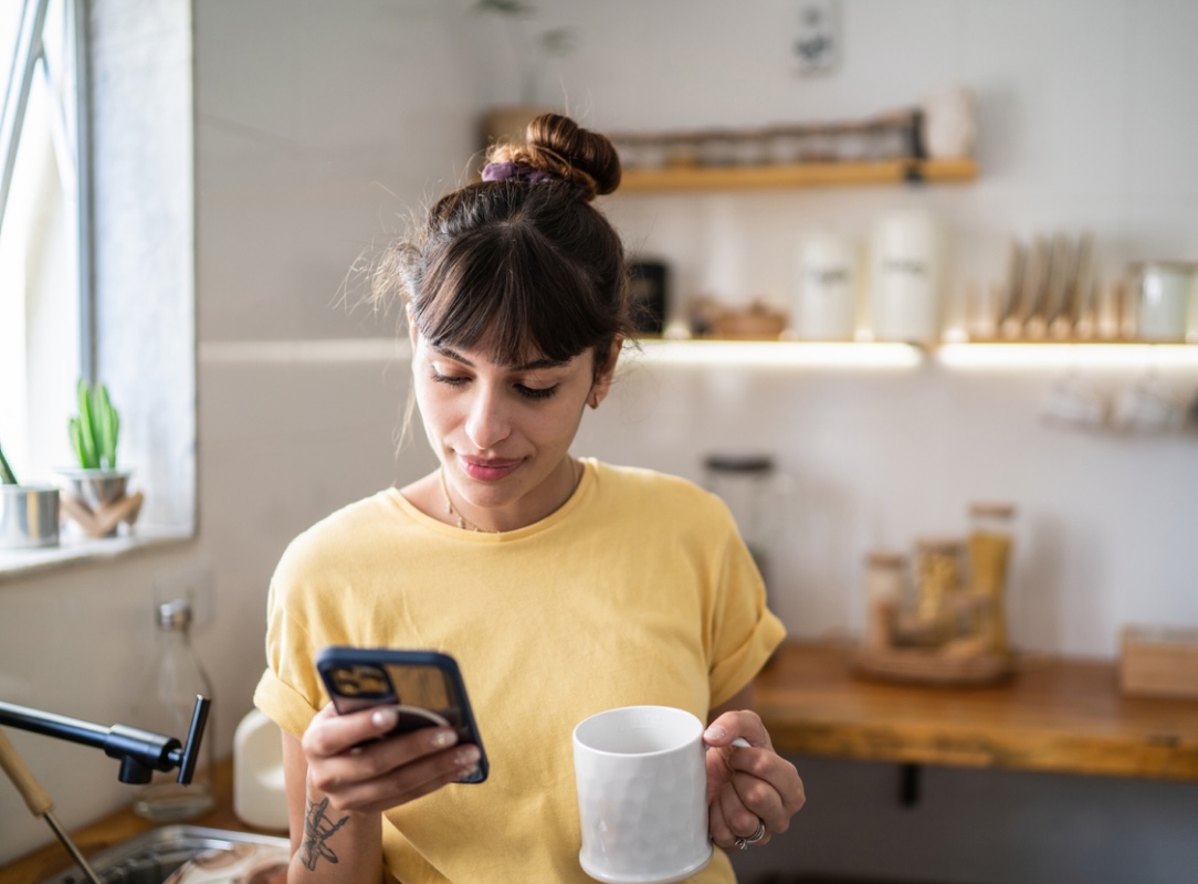 A woman in a yellow shirt holds a white mug while looking at her phone in a cozy kitchen setting.