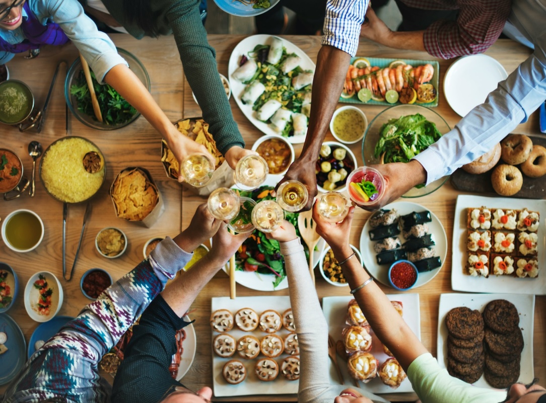 A group of hands reaching for glasses in a celebratory toast above a vast spread of diverse food.