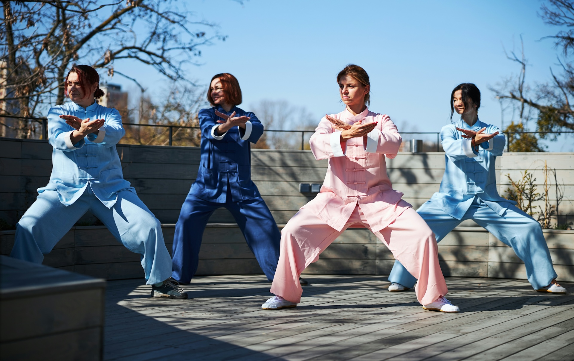 Practitioners in traditional attire performing a baduanjin qigong routine on a rooftop deck, arms crossed in the "drawing the bow" stance.
