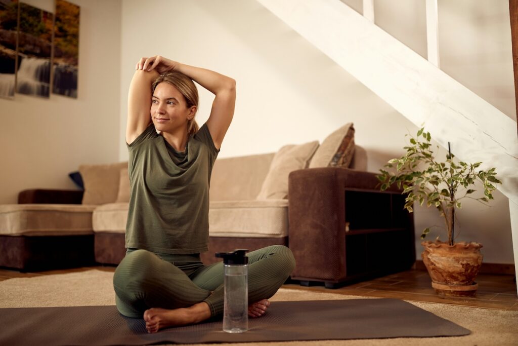 Woman sitting calmly on a yoga mat at home, stretching and looking off thoughtfully as part of practicing how to be more optimistic through mindful reflection.