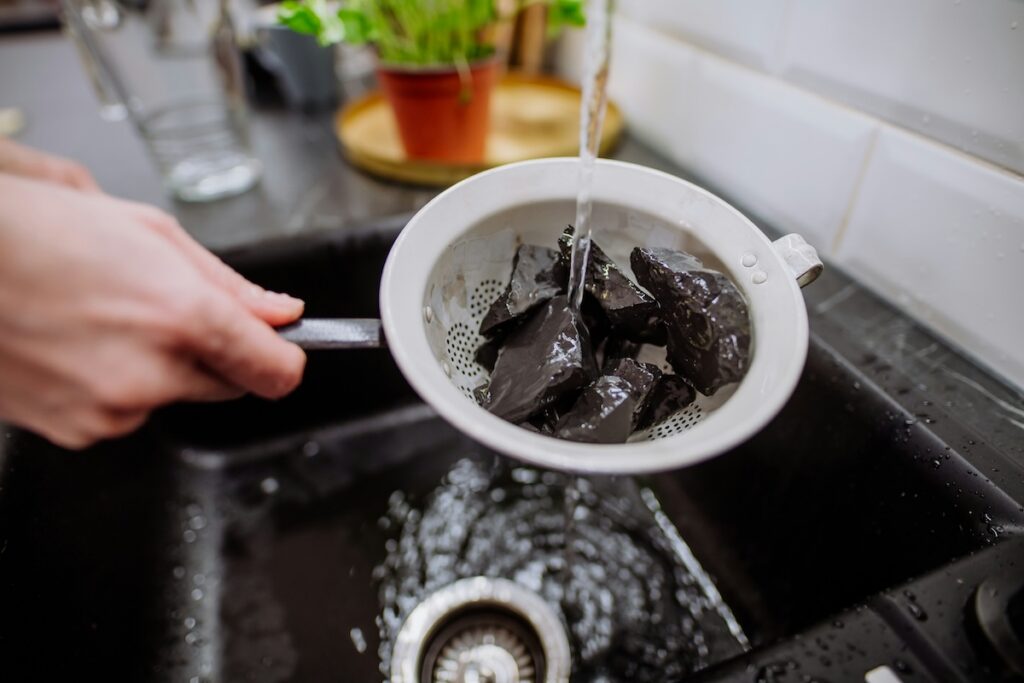 Chunks of activated carbon rinsed under running water in a strainer, illustrating a carbon filter used to remove water filters for microplastics