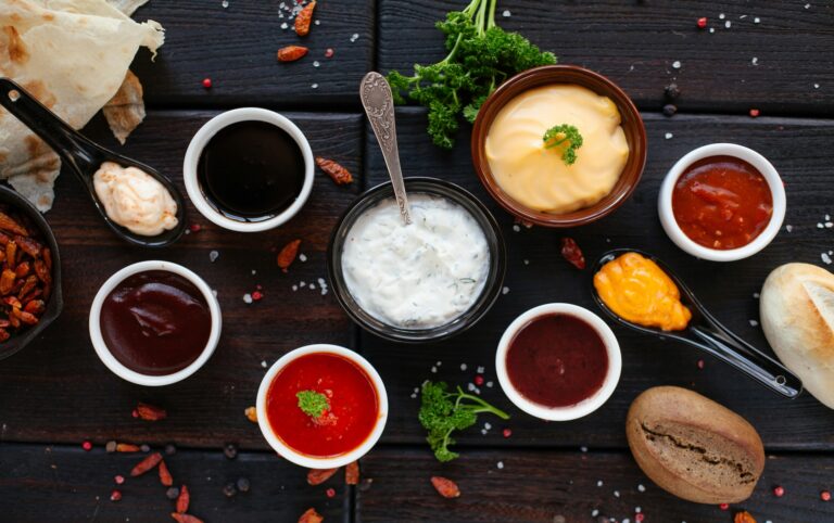 Assorted condiments for blood sugar in small bowls, including mustard, marinara, balsamic vinegar, and yogurt-based dip, arranged on a wooden table