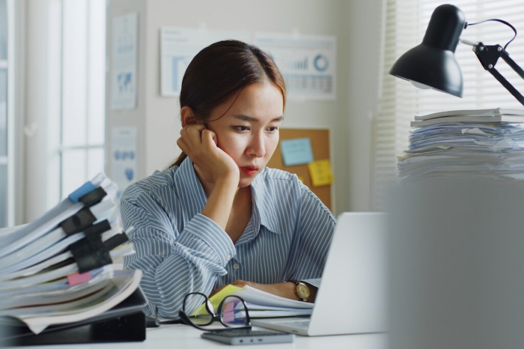 Stressed woman with hand on face at overwhelmed desk with paperwork, looking for supplements to reduce cortisol