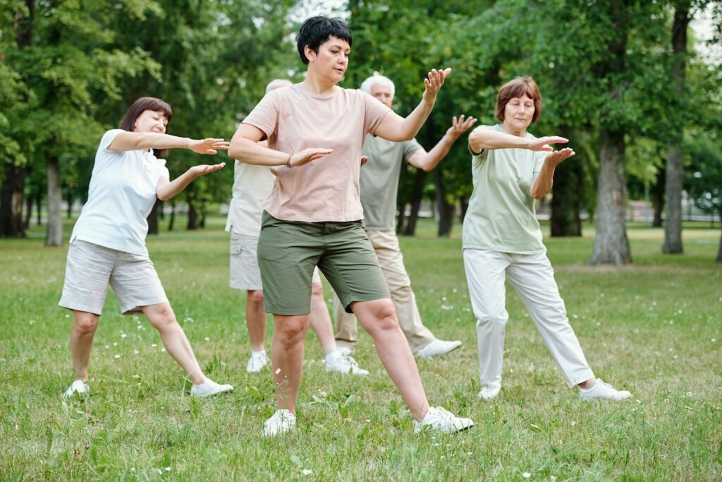Group practicing a baduanjin qigong routine outdoors, moving through the "drawing the bow" pose on a grassy park lawn.