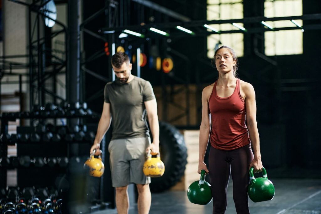 Man and woman performing farmer's carry with kettlebells to build grip strength in the gym