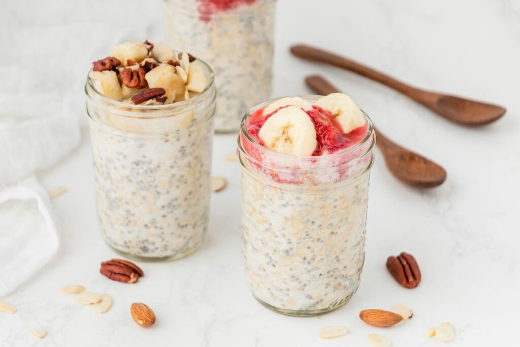 Two mason jars of high protein vegetarian meals — Greek yogurt overnight oats topped with banana, pecans, almonds, and berries