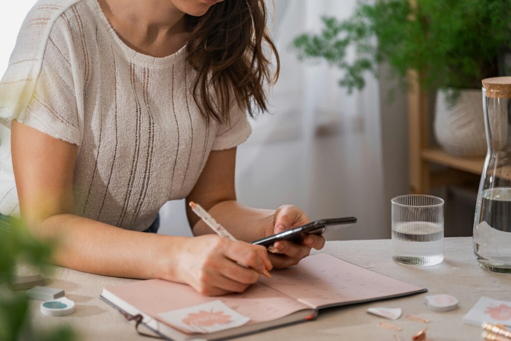 Woman writing in a planner while checking her phone, applying the WOOP goal setting method to plan her wish and action steps