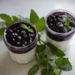 Two Greek yogurt parfait jars topped with blueberry mint jam and fresh mint leaves on a white marble counter
