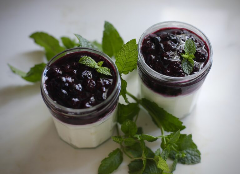 Two Greek yogurt parfait jars topped with blueberry mint jam and fresh mint leaves on a white marble counter