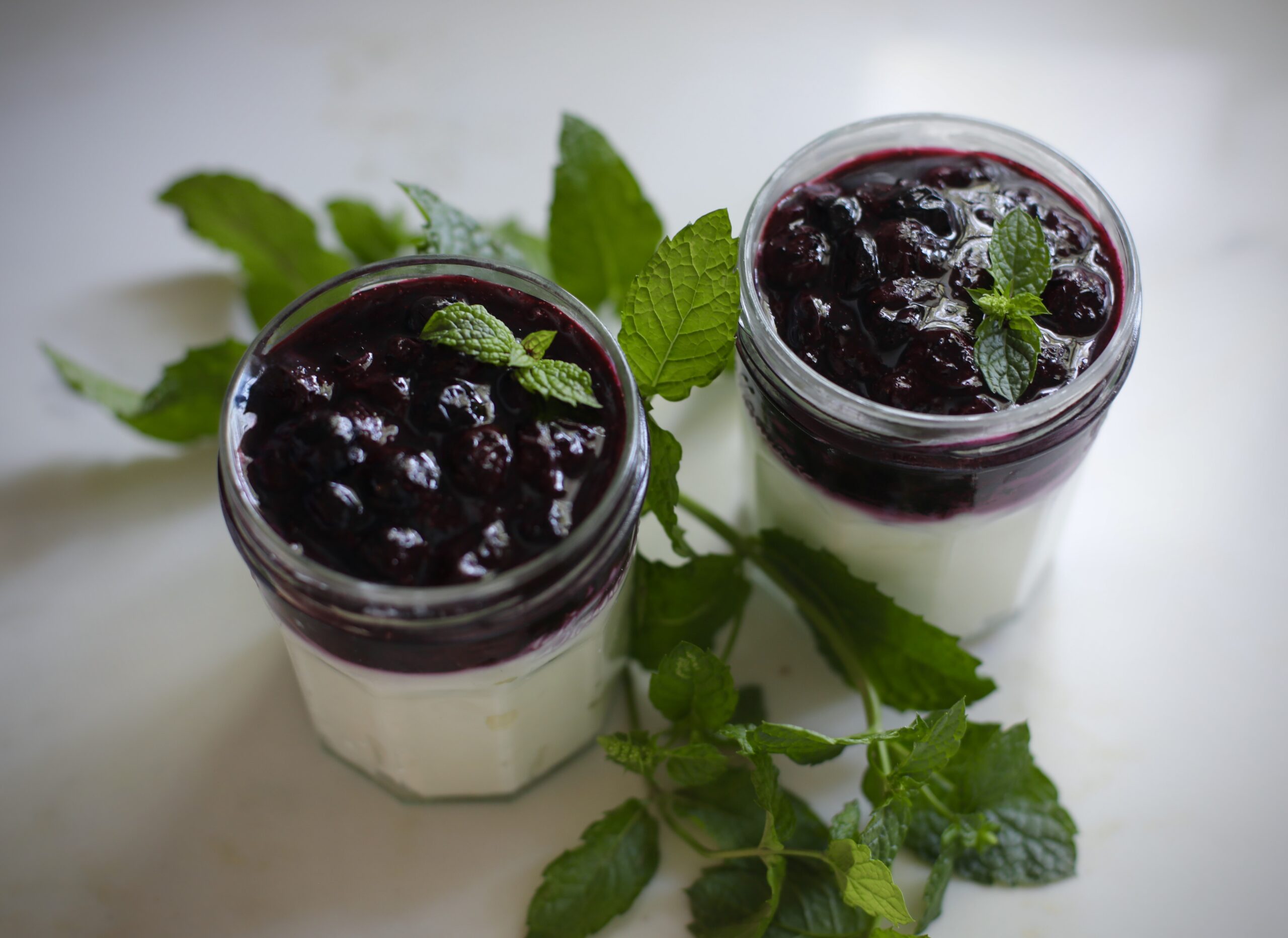 Two Greek yogurt parfait jars topped with blueberry mint jam and fresh mint leaves on a white marble counter