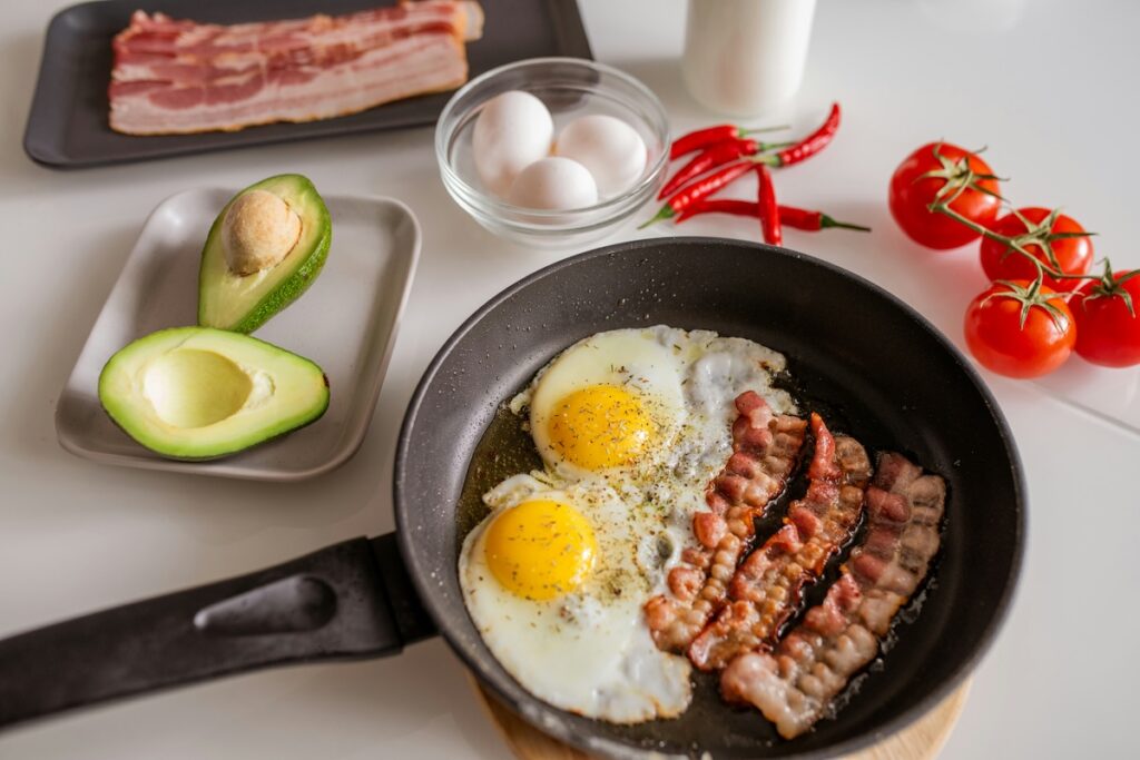 High-protein food sources including fried eggs and bacon in a skillet, with raw eggs, avocado, cherry tomatoes, and milk