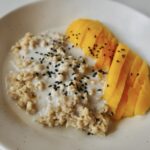 Mango sticky rice oatmeal in a bowl, topped with sliced mango, coconut sauce, and black sesame seeds