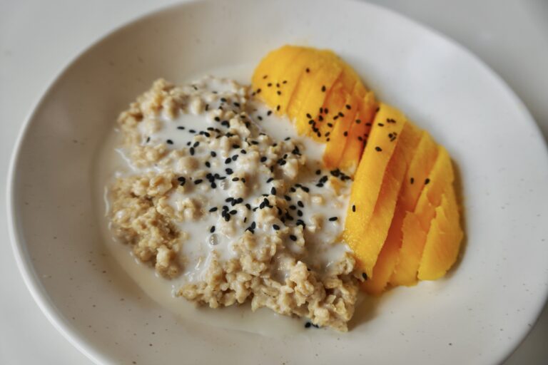 Mango sticky rice oatmeal in a bowl, topped with sliced mango, coconut sauce, and black sesame seeds