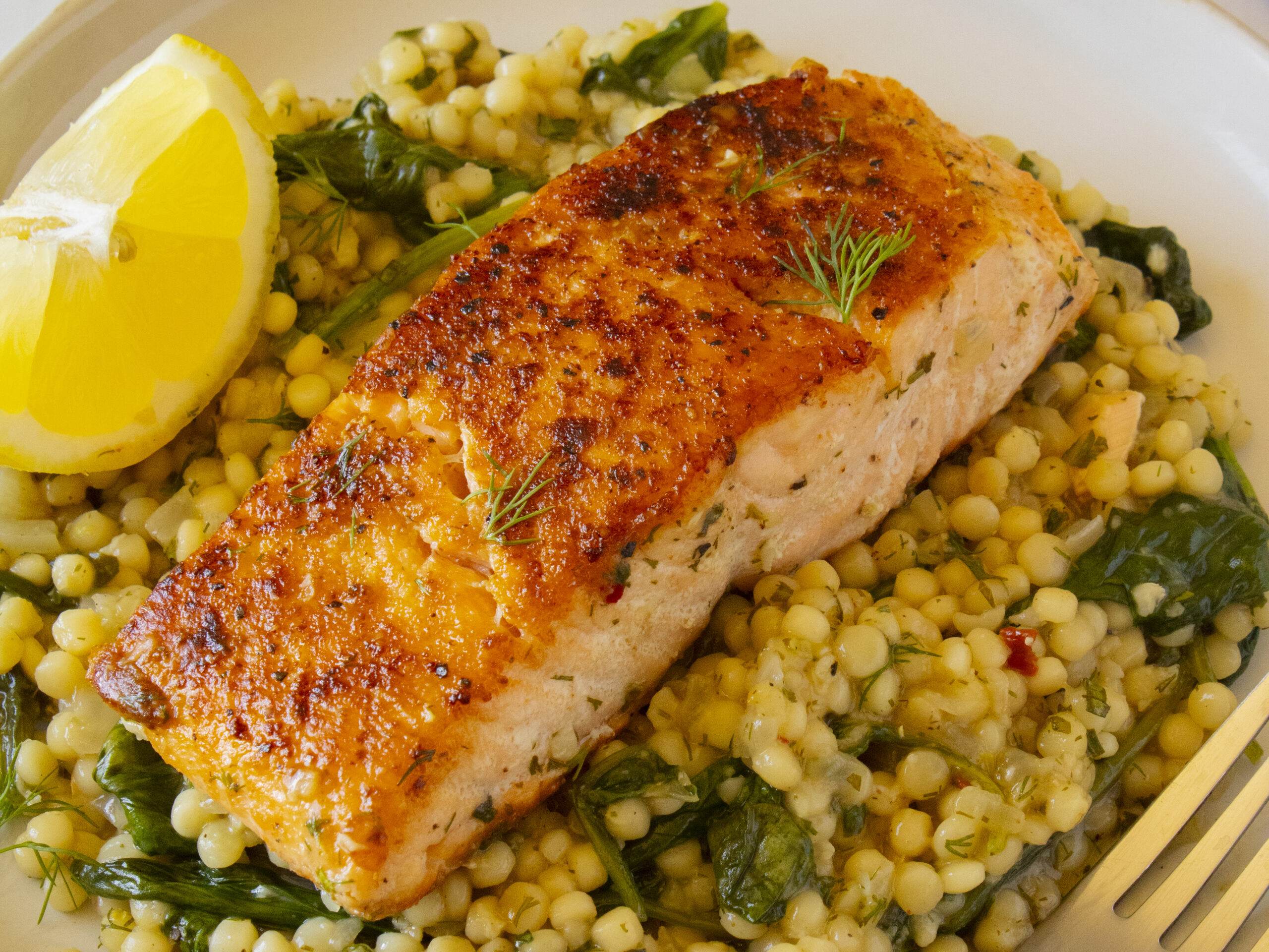 Seared herby salmon and couscous with spinach, fresh dill, and a lemon wedge on a white plate