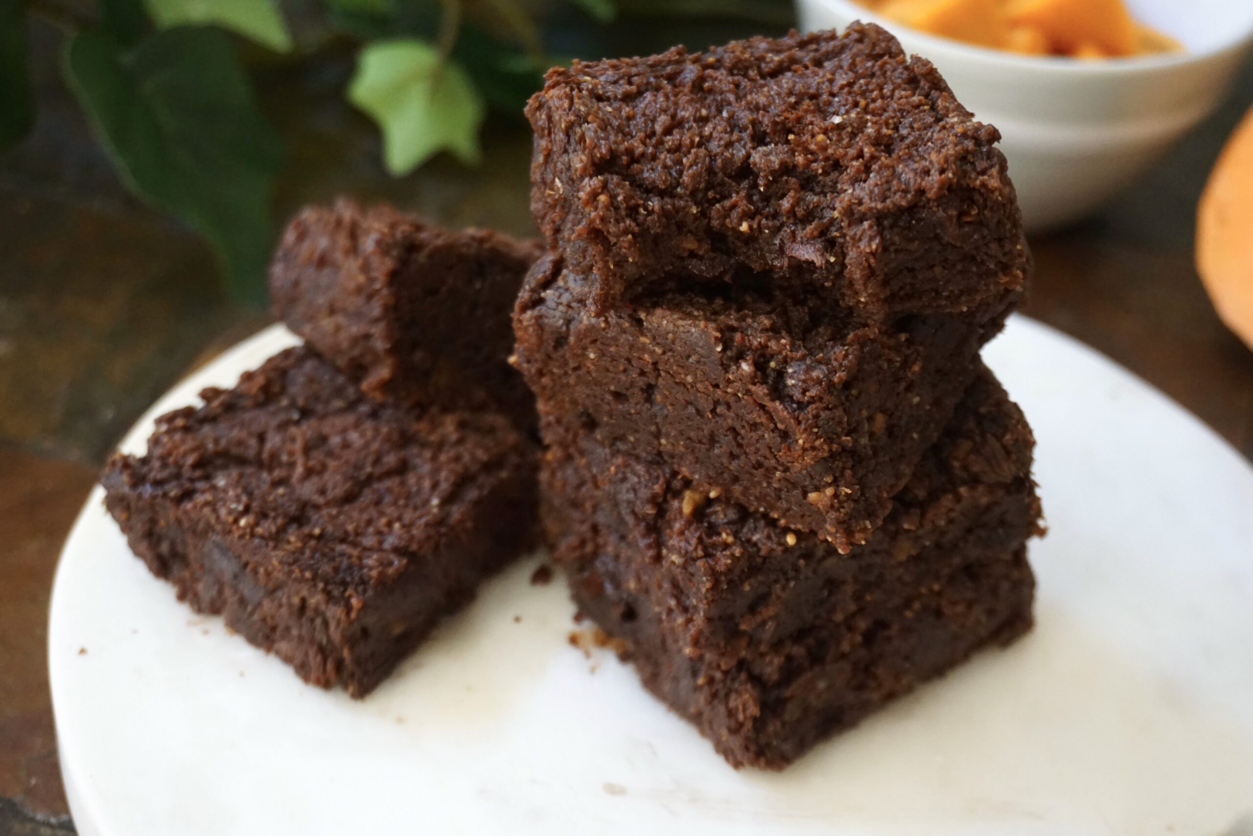 Stacked squares of fudgy sweet potato brownies on a white plate, with roasted sweet potato in the background.