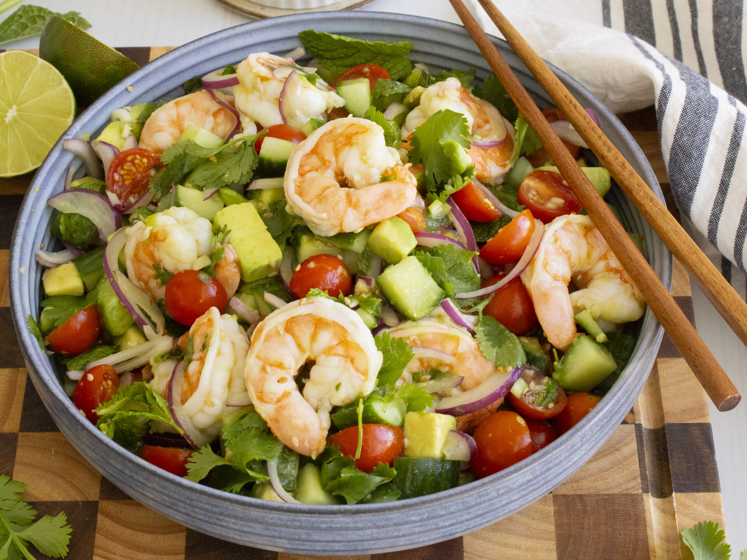 Vietnamese shrimp salad with poached shrimp, avocado, cucumber, cherry tomatoes, red onion, and fresh herbs in a blue bowl with chopsticks and lime