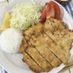 Sliced air fryer chicken katsu on a plate with rice, shredded cabbage, tomato, and a bowl of creamy wasabi yogurt sauce.