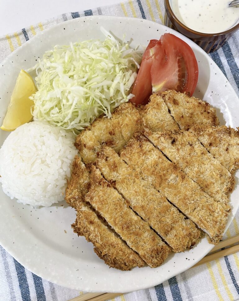 Sliced air fryer chicken katsu on a plate with rice, shredded cabbage, tomato, and a bowl of creamy wasabi yogurt sauce.