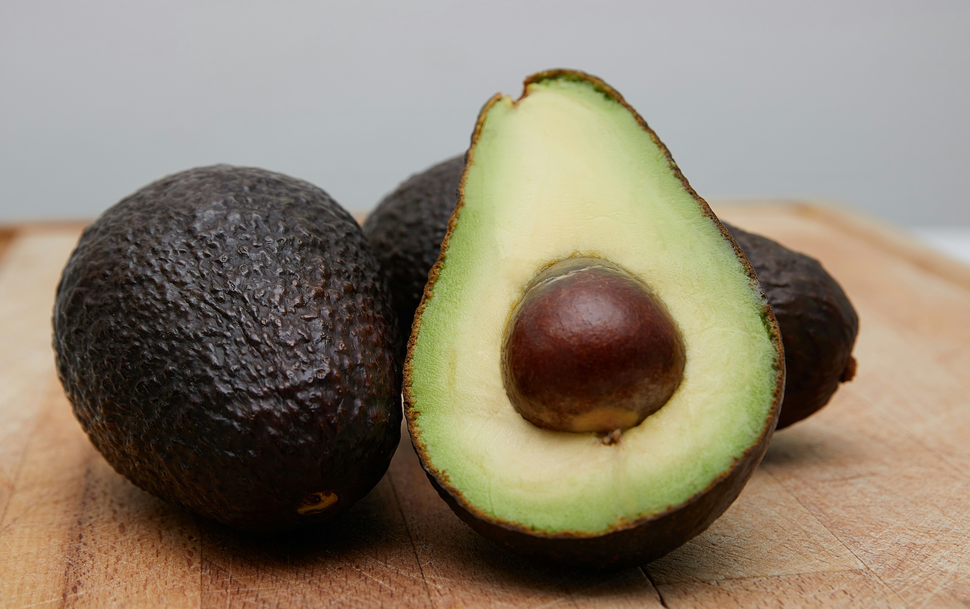 A ripe avocado half with its pit beside whole avocados on a wooden table.