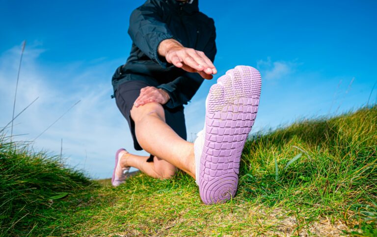 Person kneeling outdoors and extending a leg wearing zero-drop toe shoes, incorporating foot strengthening exercises into an outdoor stretching routine