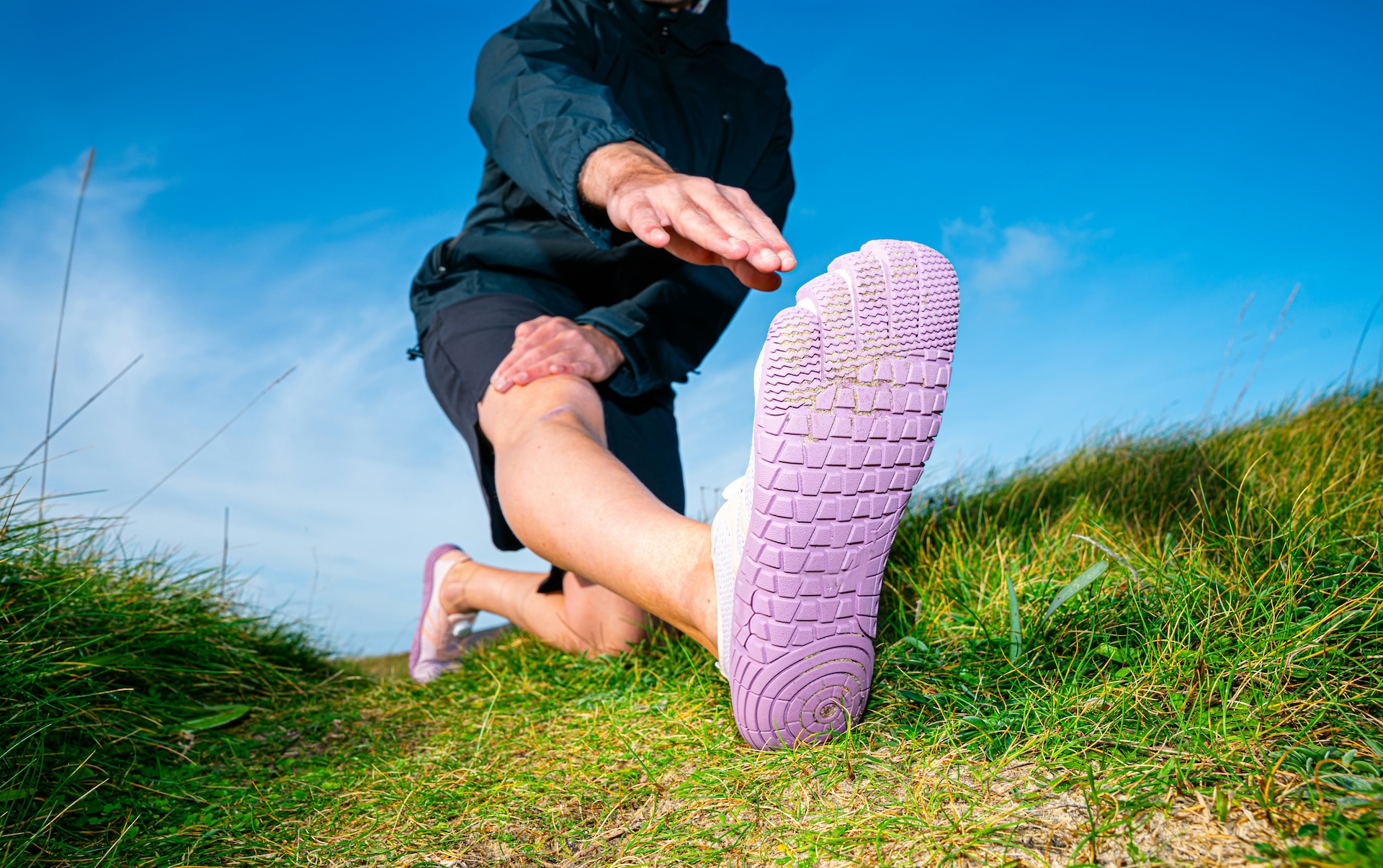 Person kneeling outdoors and extending a leg wearing zero-drop toe shoes, incorporating foot strengthening exercises into an outdoor stretching routine