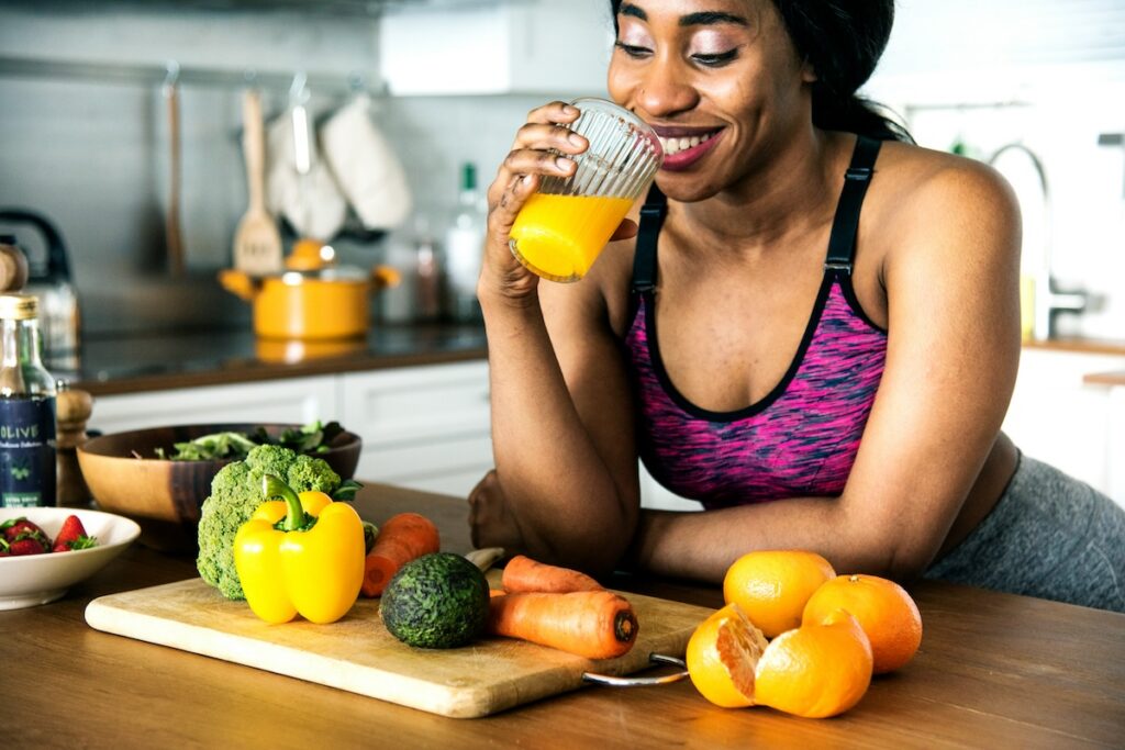 Woman drinking a glass of orange juice in a bright kitchen with fresh fruit and vegetables on the counter.