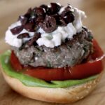 Lamb burger recipe served open-faced on a brioche bun with butter lettuce, tomato, feta yogurt sauce, and kalamata olive spread
