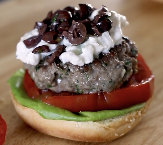 Lamb burger recipe served open-faced on a brioche bun with butter lettuce, tomato, feta yogurt sauce, and kalamata olive spread