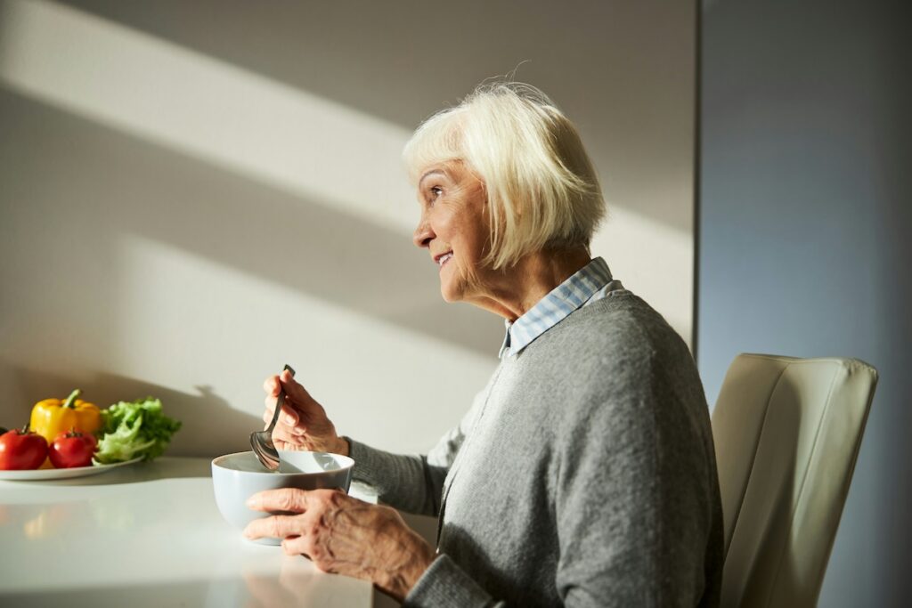 Older woman smiling while eating a bowl of food at her table, practicing intermittent fasting for women over 50 with a nutrient-dense meal during her eating window