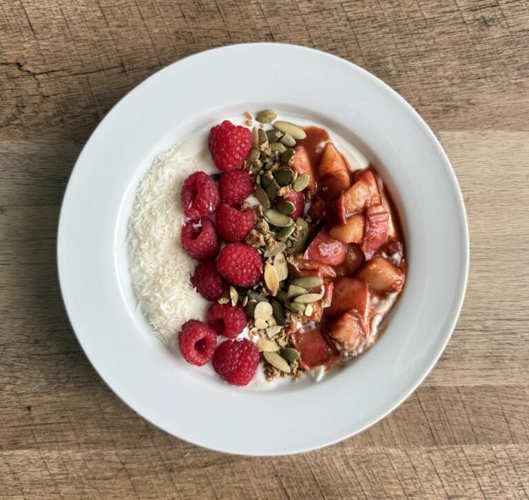 Overhead view of a peach compote yogurt bowl with Greek yogurt, raspberries, shredded coconut, and pumpkin seeds on a white plate.