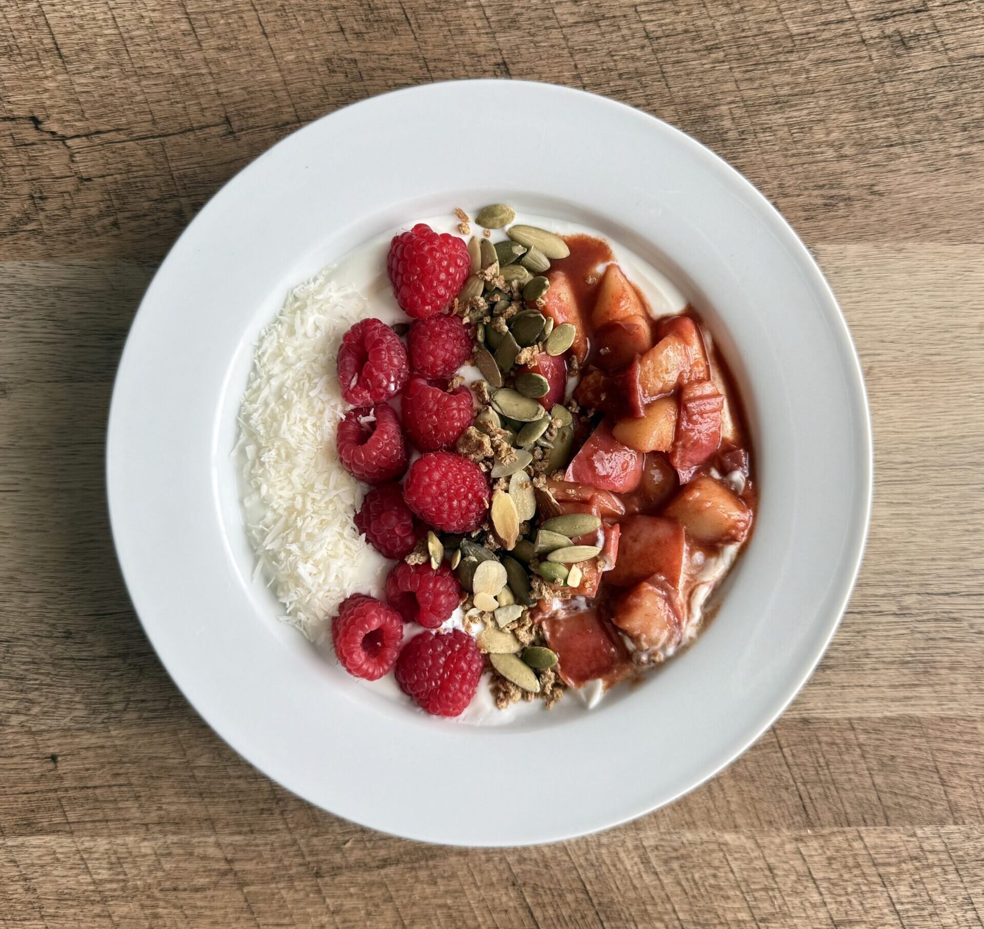 Overhead view of a peach compote yogurt bowl with Greek yogurt, raspberries, shredded coconut, and pumpkin seeds on a white plate.