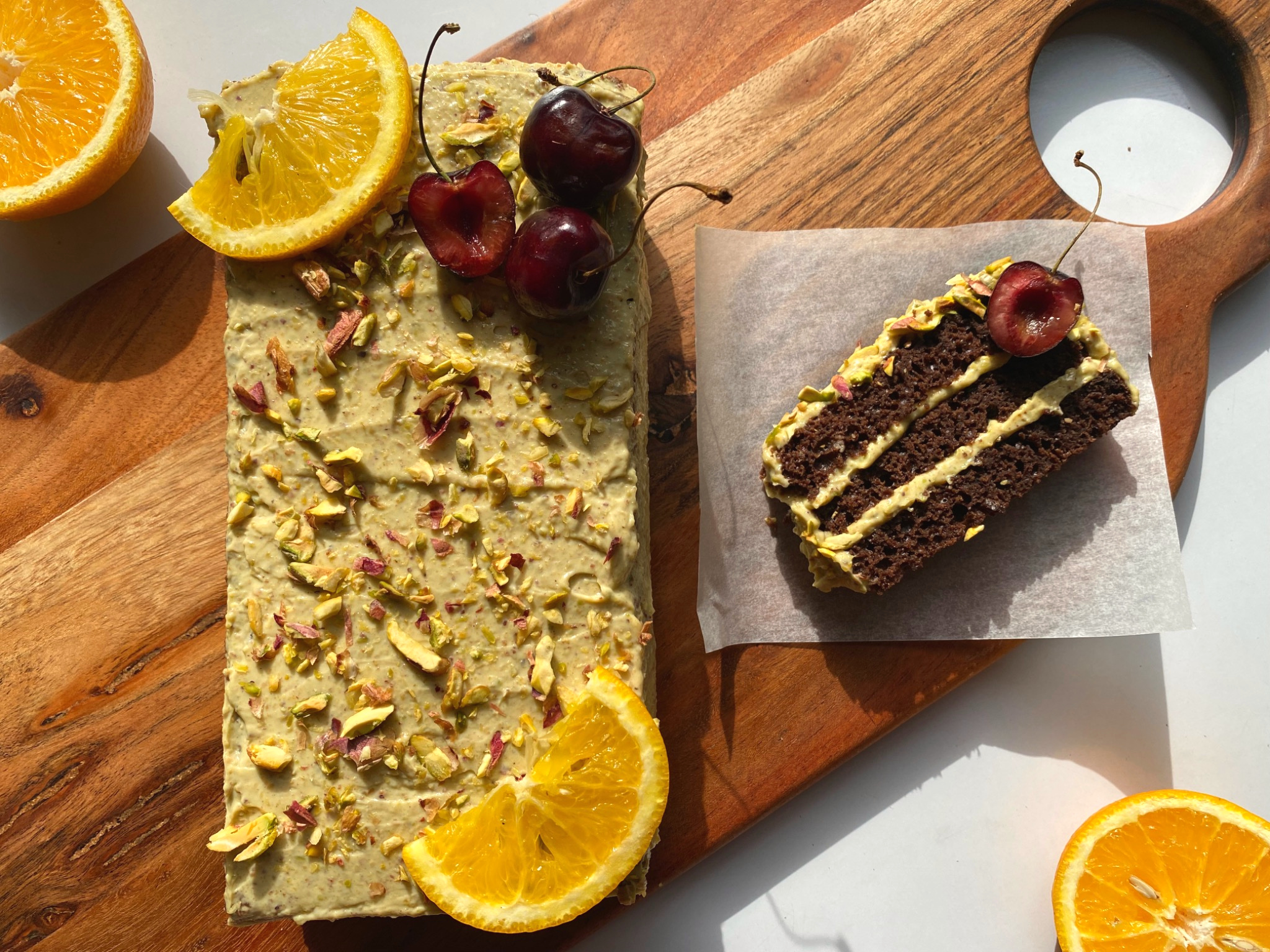 Chocolate pistachio cake topped with chopped pistachios, orange slices, and cherries, with a sliced piece showing three layers of pistachio yogurt cream