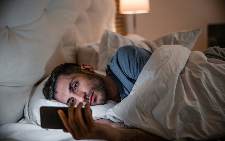A man lies awake in bed scrolling on his phone late at night, a common pattern in ADHD and sleep problems.