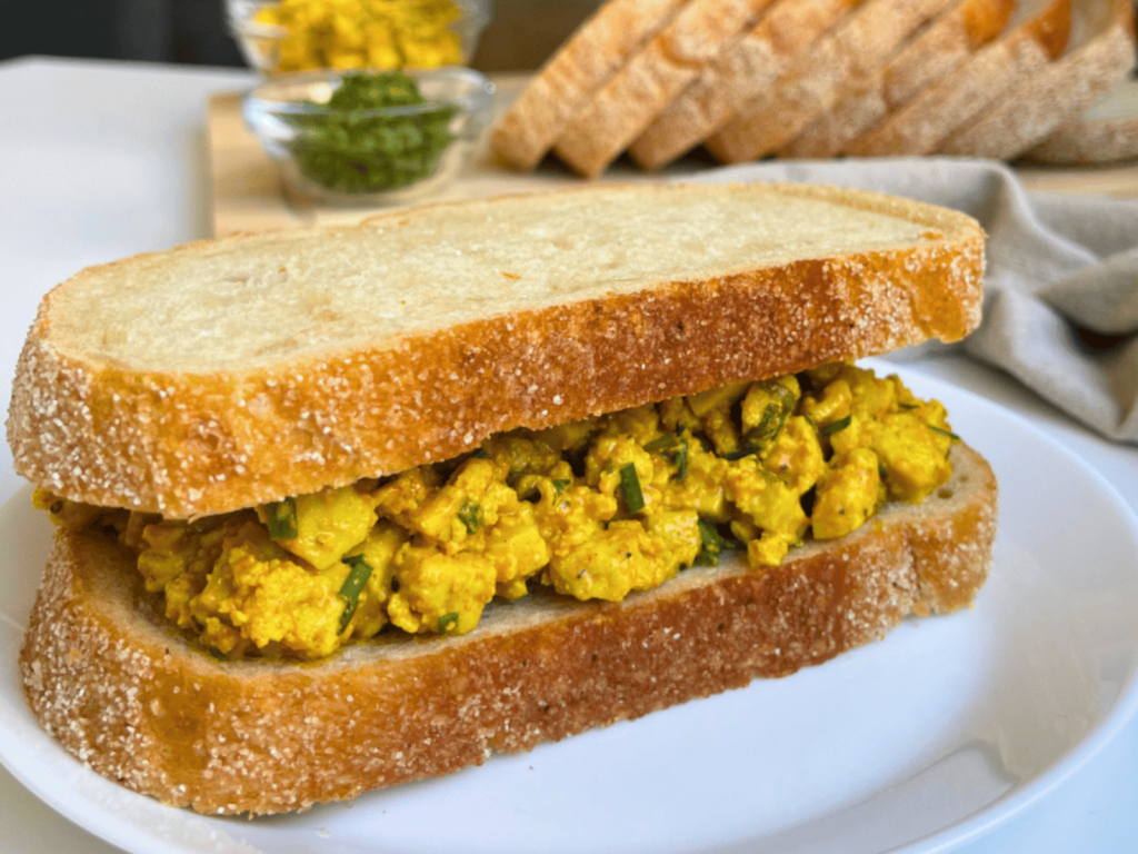 Vegan egg salad sandwich on crusty bread with smashed turmeric tofu and chives, a high-protein meal prep lunch