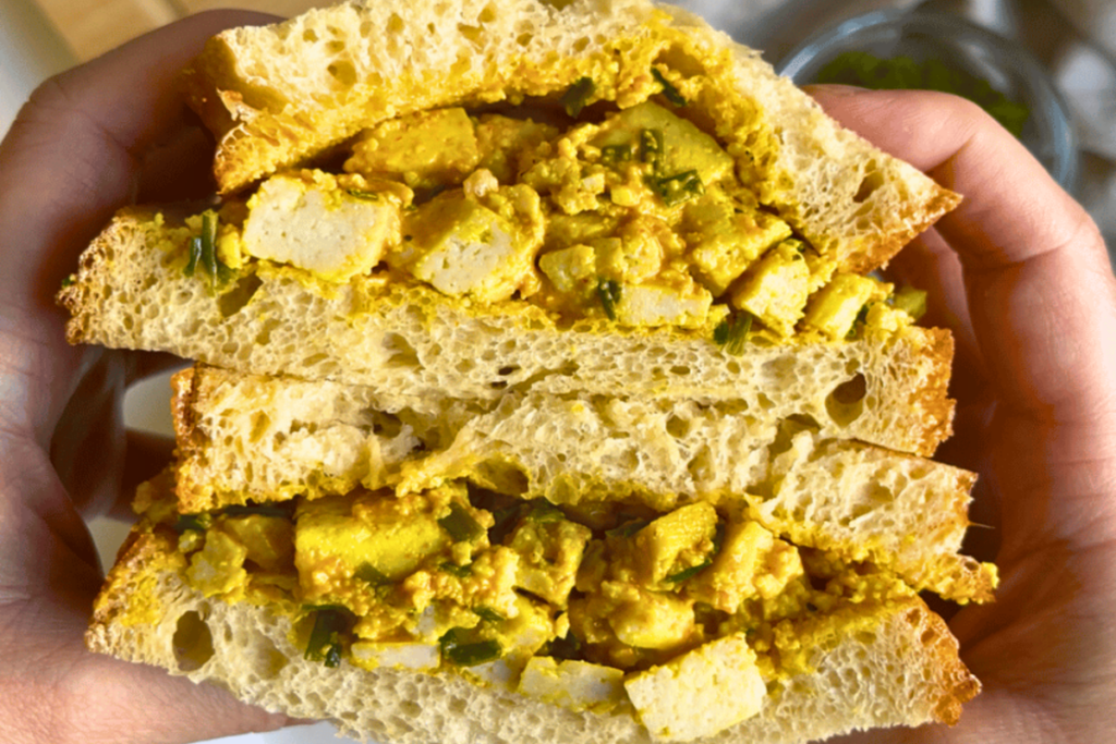 High protein meal prep lunch: hands holding a vegan egg salad sandwich filled with turmeric tofu and chives