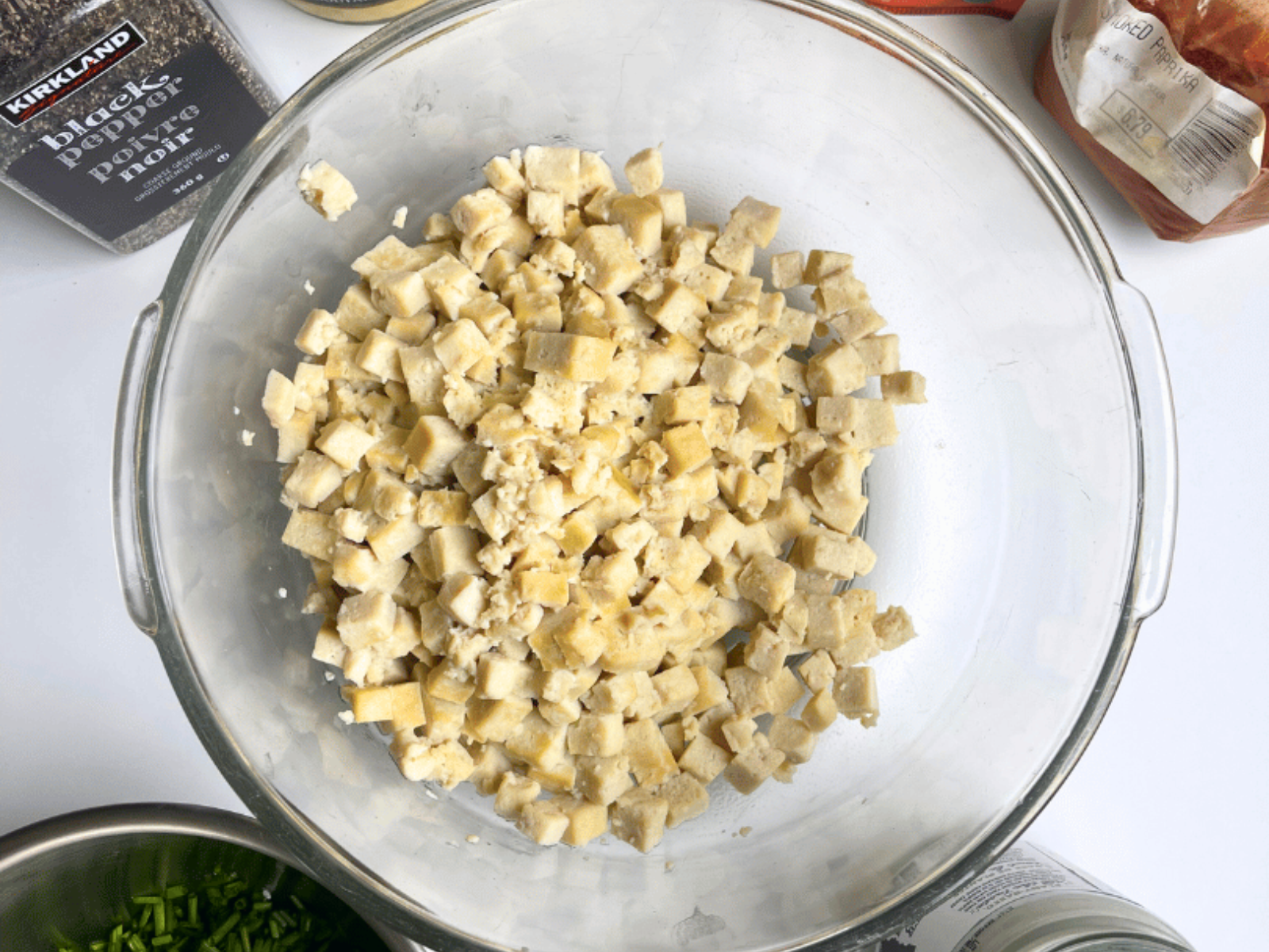 Boiled tofu cubes in a glass bowl, the first step in making vegan egg salad, with spices and chives nearby