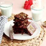 Stack of healthy brownies topped with salted pumpkin seeds on a white plate, with two glasses of milk in the background