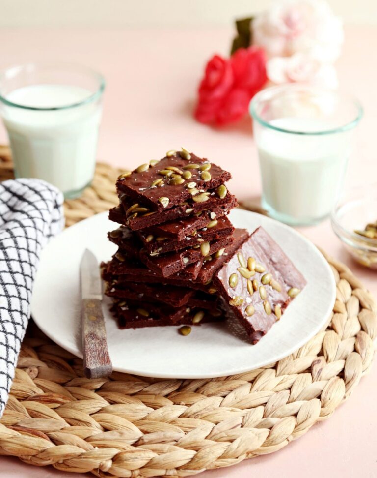 Stack of healthy brownies topped with salted pumpkin seeds on a white plate, with two glasses of milk in the background