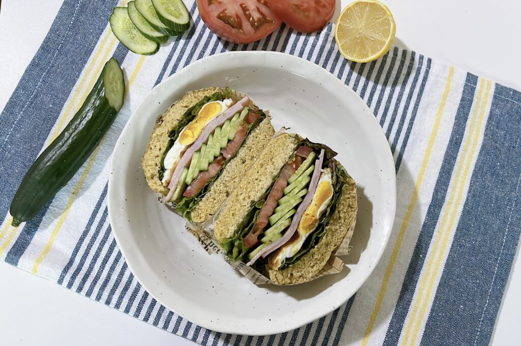 Gluten-free breakfast sandwich on oat flour bread, cut in half and filled with ham, fried egg, tomato, cucumber, and lettuce