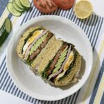 Gluten-free breakfast sandwich on oat flour bread, cut in half and filled with ham, fried egg, tomato, cucumber, and lettuce