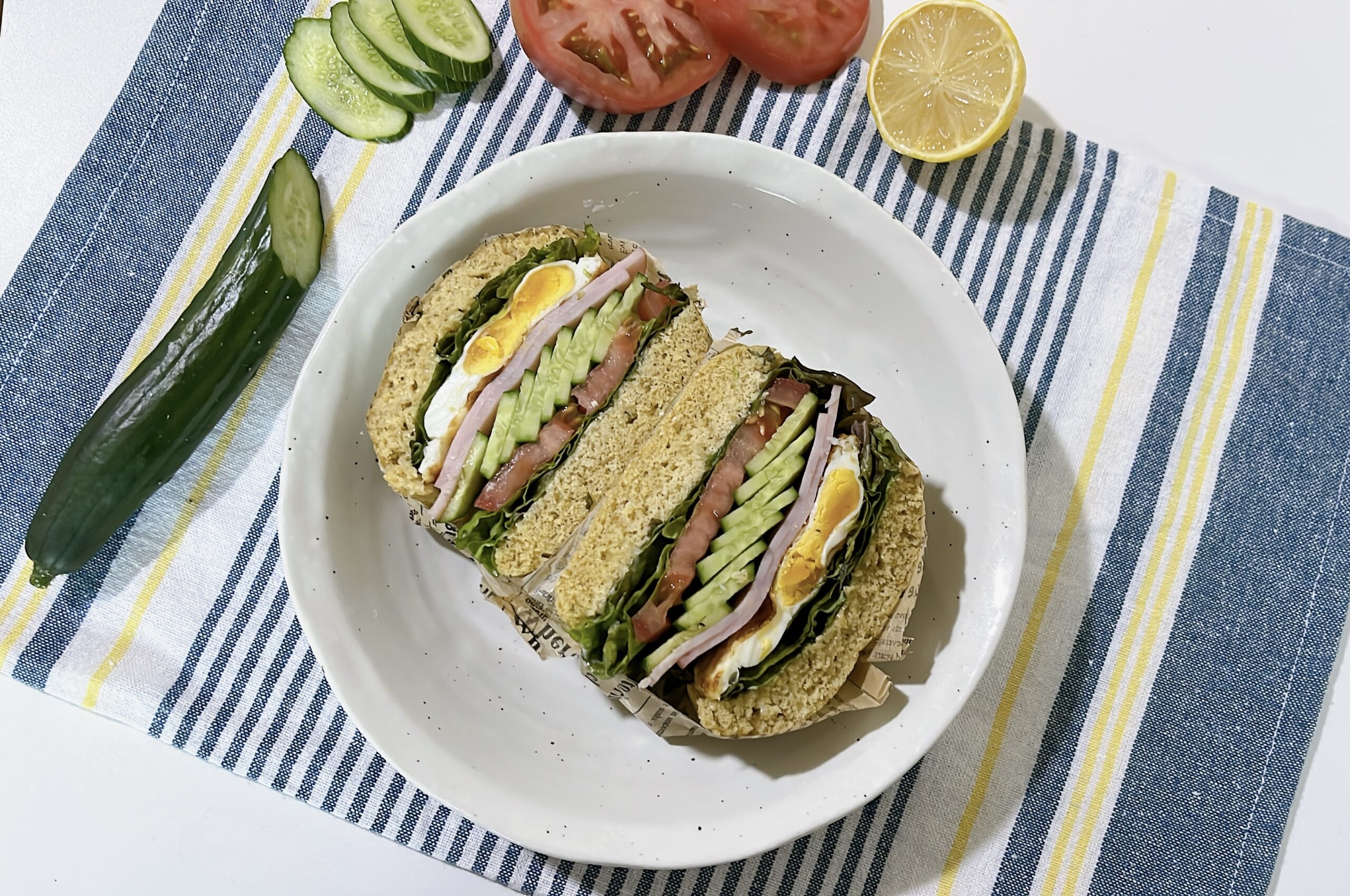 Gluten-free breakfast sandwich on oat flour bread, cut in half and filled with ham, fried egg, tomato, cucumber, and lettuce