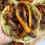 Hand holding steak fajitas with avocado sauce, sliced beef, and red and yellow bell peppers on a flour tortilla