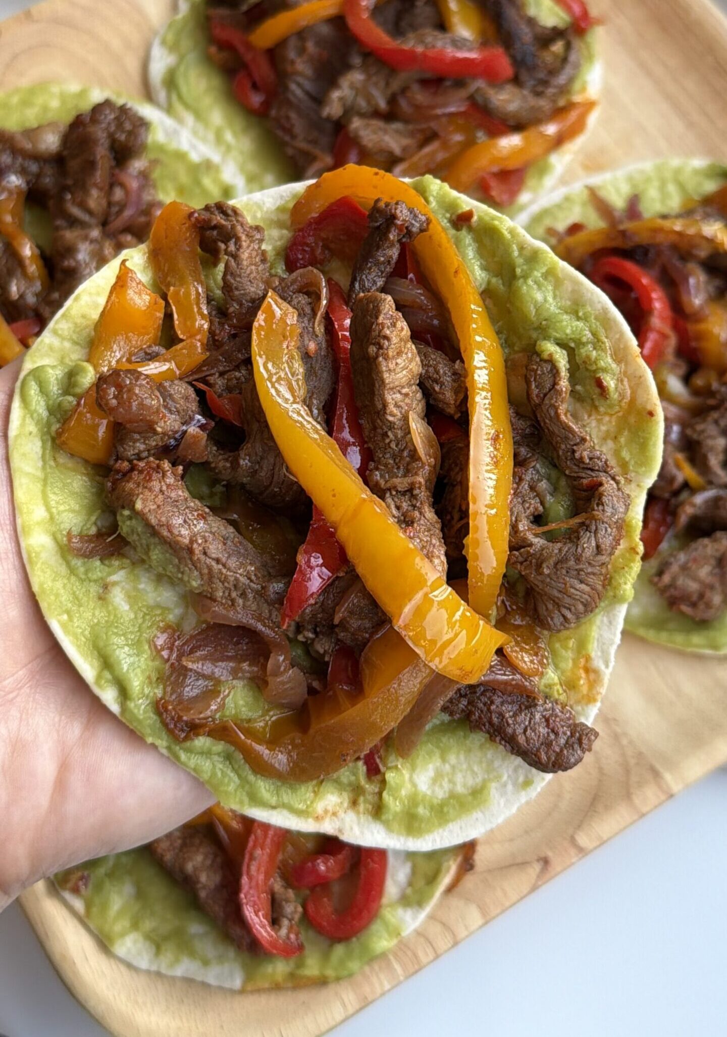 Hand holding steak fajitas with avocado sauce, sliced beef, and red and yellow bell peppers on a flour tortilla