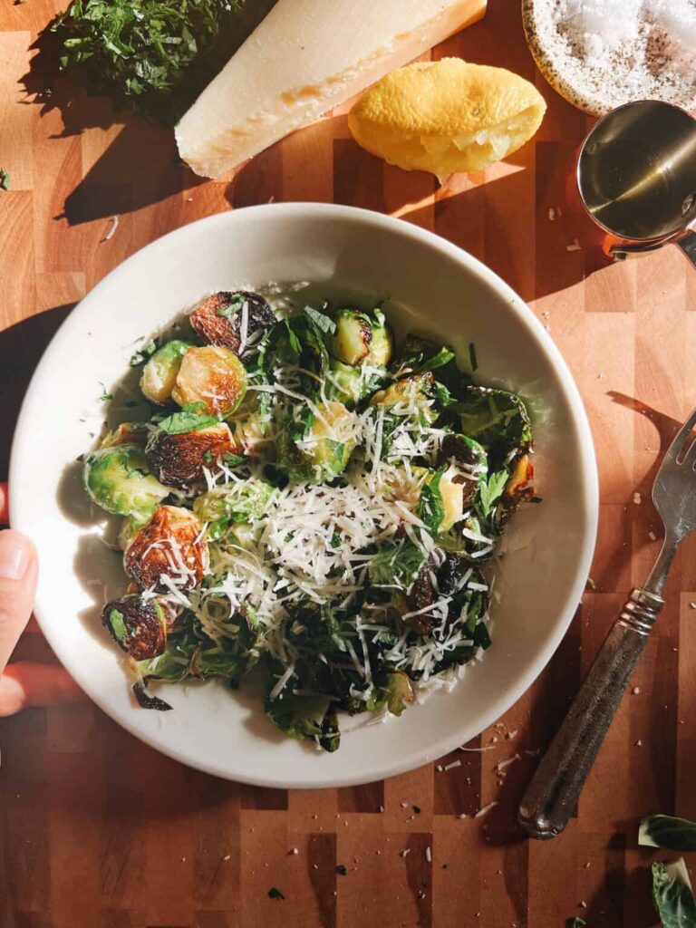 Bowl of crispy Brussels sprouts with golden cut sides, topped with grated Parmesan and chopped parsley, next to a lemon half, a wedge of Parmesan, and olive oil on a wooden board